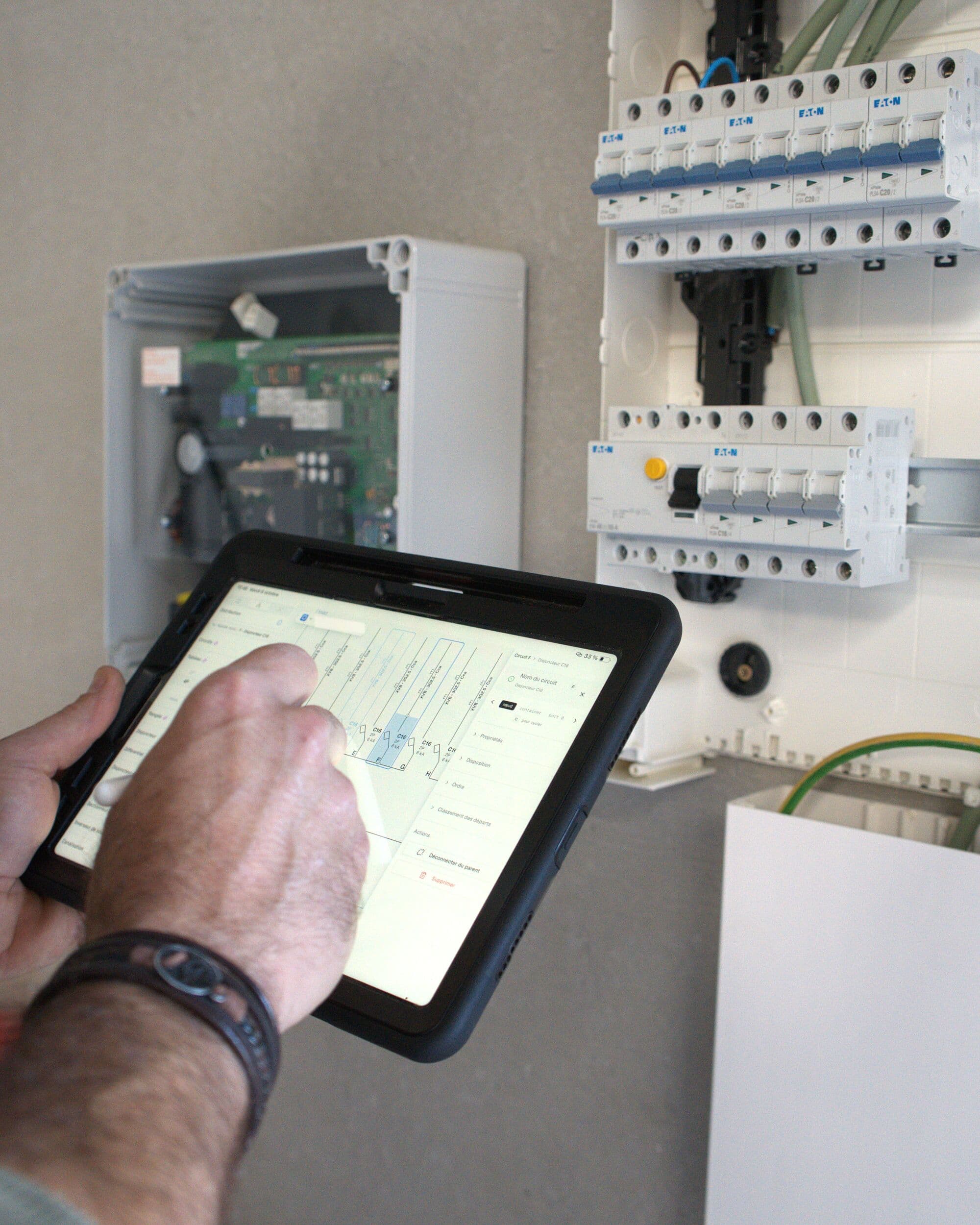 Tablette en main devant un tableau électrique, schéma unifilaire ElectriCAD à l'écran